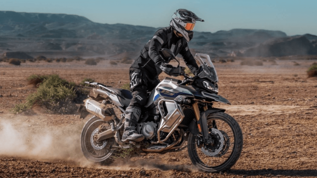Motociclista conduciendo una moto de aventura en un paisaje desértico con montañas al fondo.