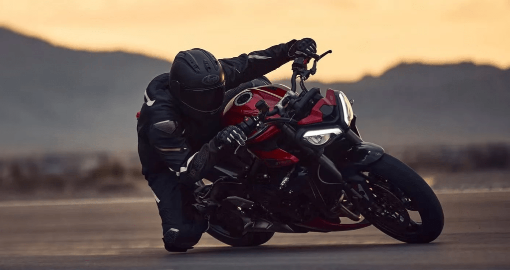 Un piloto de motocicleta vestido de negro inclinado sobre una moto roja durante una curva en un paisaje montañoso al atardecer.