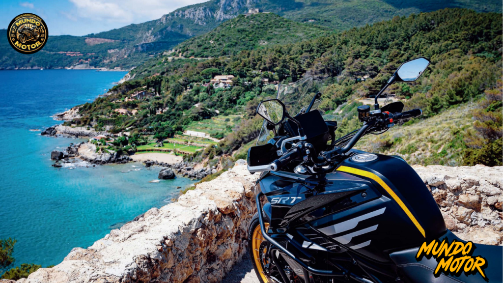 Vista lateral de la motocicleta QJMotor SRT 900 en un paisaje costero, con montañas al fondo y un mar azul brillante.