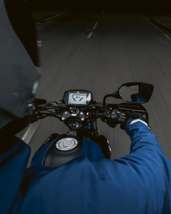 Vista desde el manillar de una motocicleta en movimiento, mostrando el velocímetro digital con una lectura de 24 km/h, y un conductor con chaqueta azul y casco.