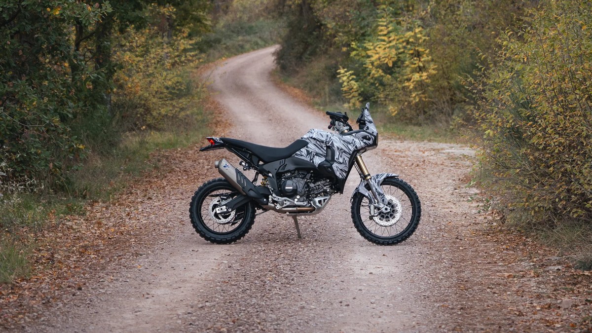 Motocicleta Ducati DesertX 2026 en una carretera de tierra, equipada para conducción off-road, con un diseño moderno y una estética en camuflaje.