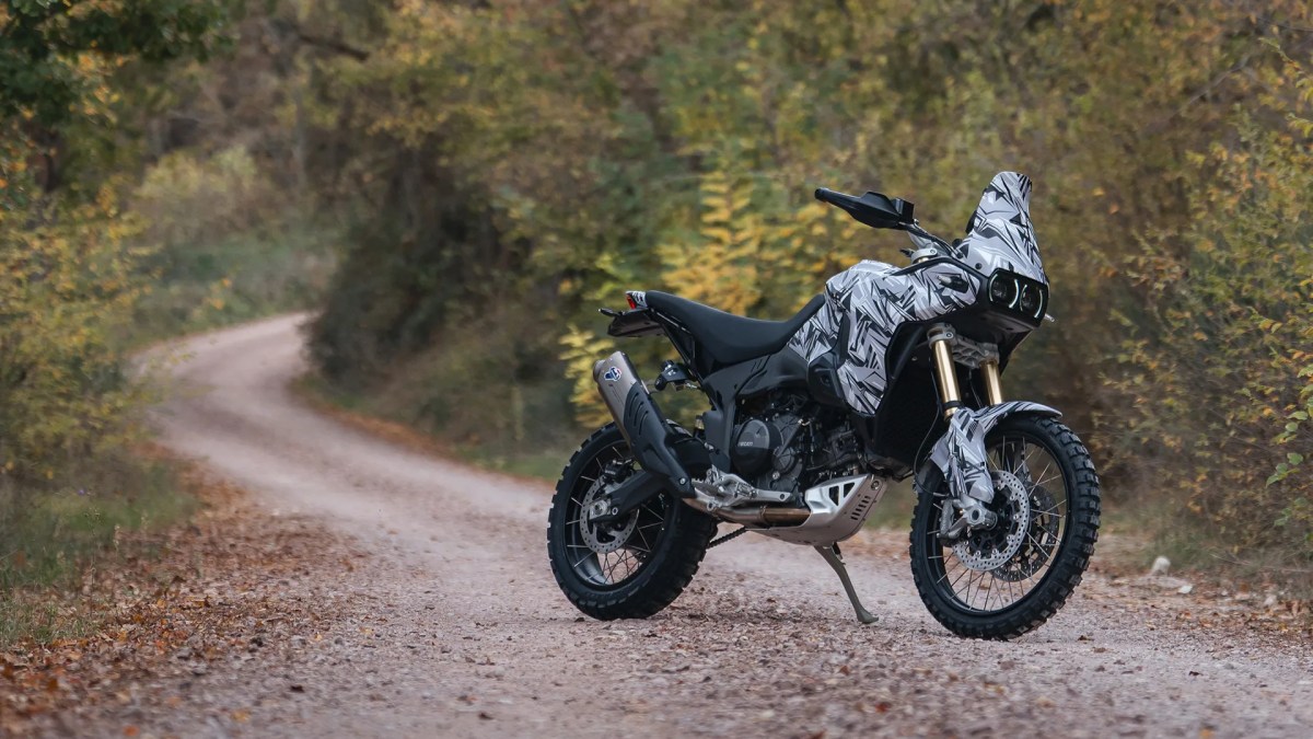 Ducati DesertX 2026 en un camino de tierra rodeado de árboles, con un diseño de camuflaje.