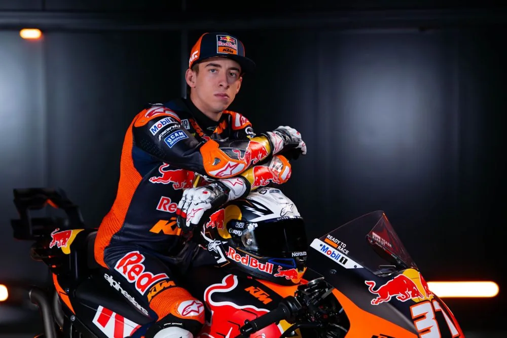 Piloto de MotoGP posando junto a su moto de la escudería Red Bull KTM, luciendo el equipo y una expresión seria en un entorno oscuro.