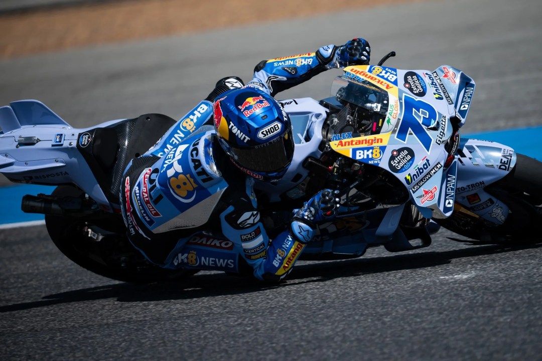 Un piloto de motociclismo en una curva, montando una moto de carreras decorada con patrocinadores y números en su vestido. El fondo muestra una pista de carreras.