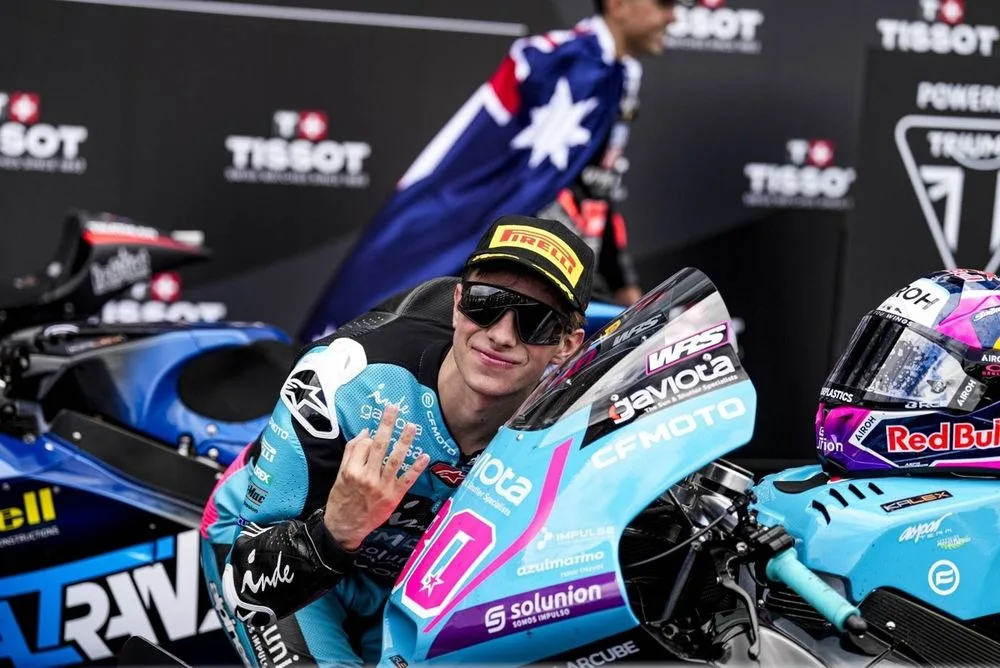 Piloto con gafas de sol posando al lado de su moto de competición, mostrando el gesto de la victoria con una mano, en un ambiente de celebración.