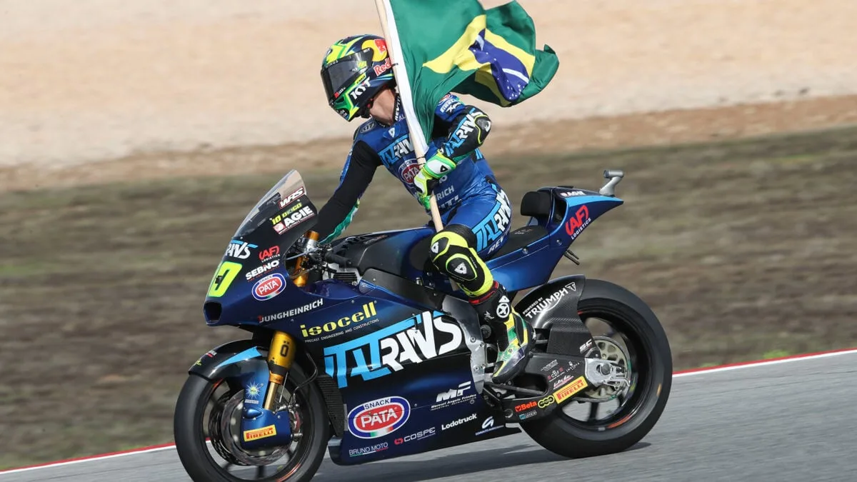 Piloto de Moto2 celebrando su victoria mientras sostiene la bandera de Brasil, montado sobre su moto en la pista.