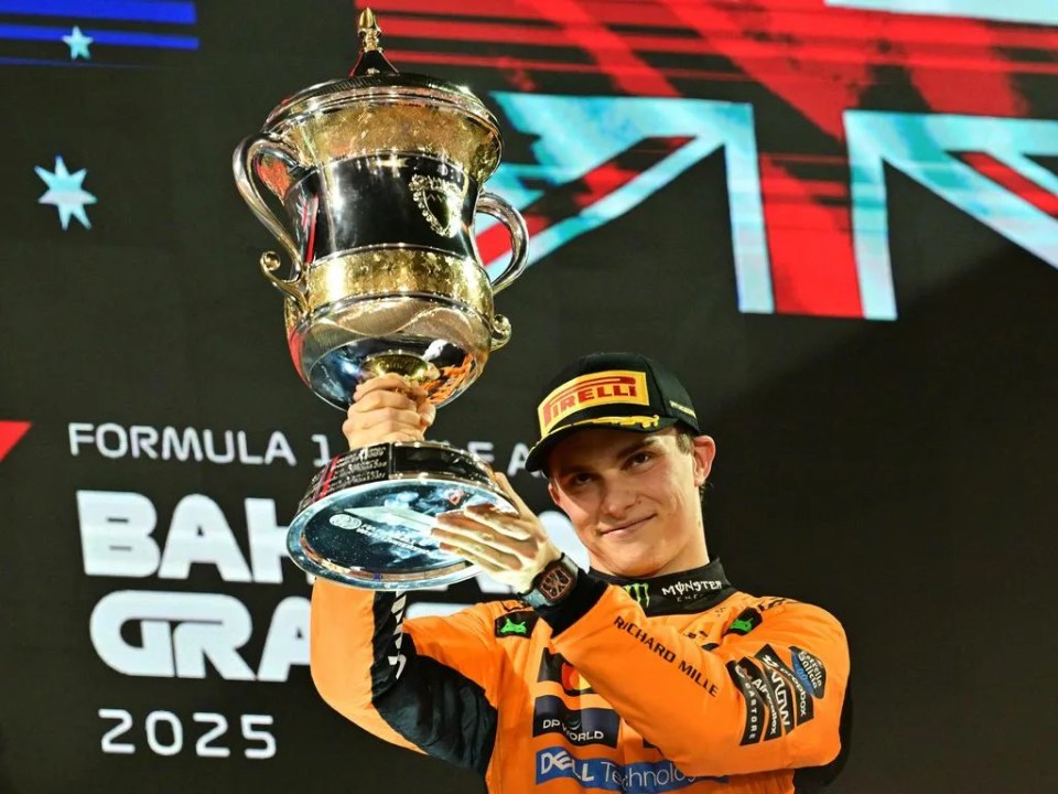 Un piloto de Fórmula 1 sostiene un trofeo en la ceremonia de premiación, con una bandera de fondo.