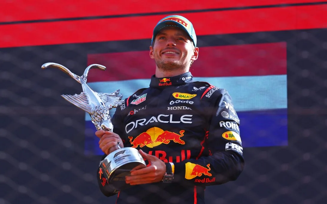 Piloto sosteniendo un trofeo en el podio, con el logo de Red Bull en su traje y fondo que muestra una bandera decorativa.