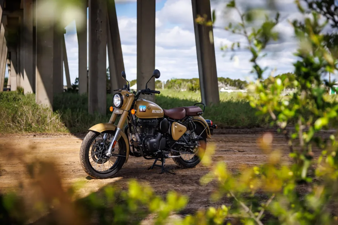 Motocicleta Royal Enfield Meteor 350 de color beige en un entorno natural con vegetación y un puente de fondo.