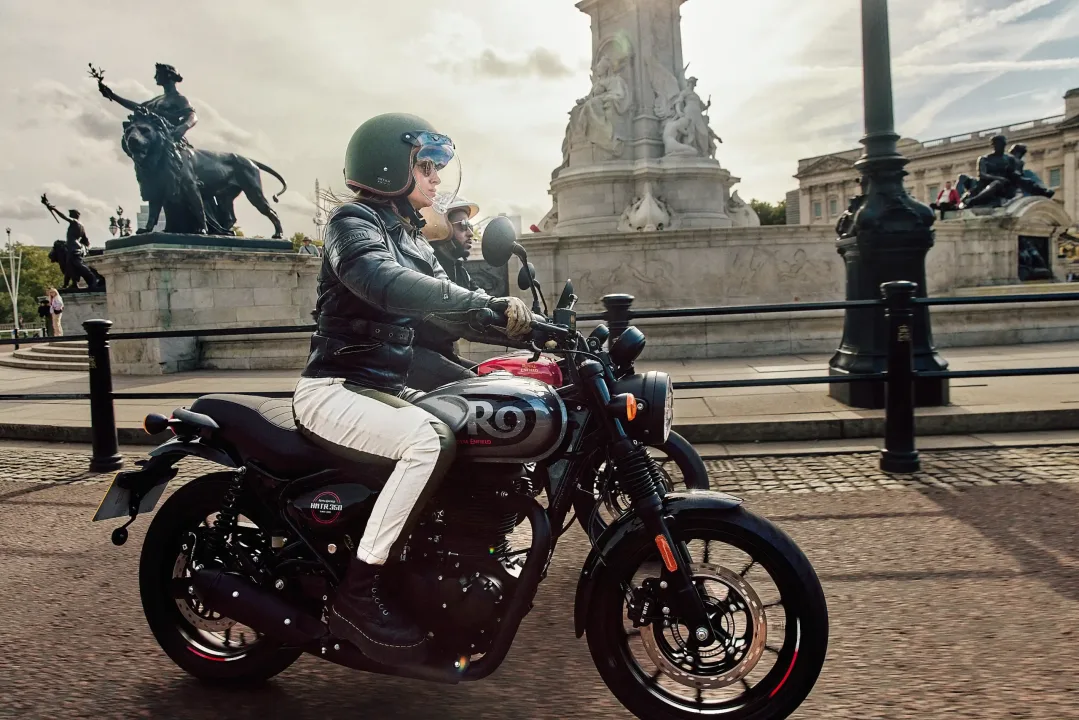 Una motocicleta Royal Enfield HNTR 350 en movimiento, pilotada por una mujer con casco, pasando por un entorno urbano con monumentos al fondo.