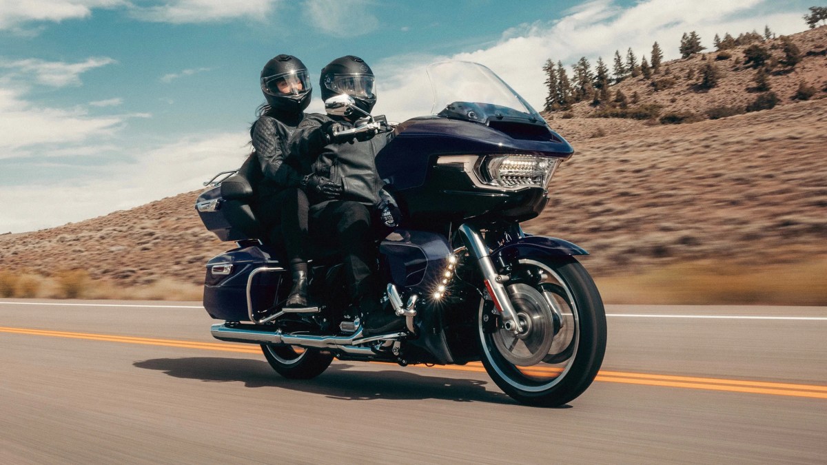 Two riders wearing helmets on a blue Harley-Davidson Road Glide Limited 2026 motorcycle, cruising on a highway with a mountainous landscape in the background.