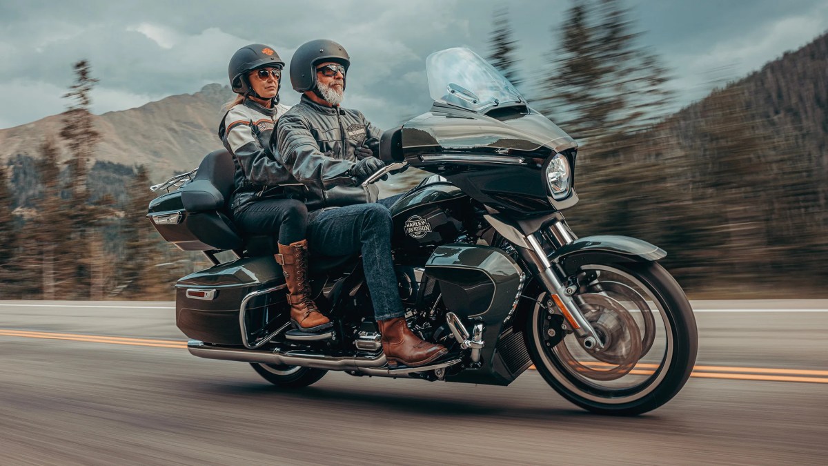 A couple riding a motorcycle on a scenic road, wearing helmets and leather jackets, with mountains in the background.