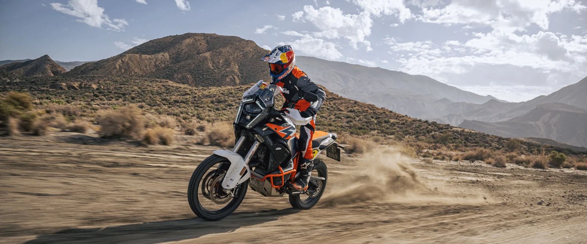 A motorcyclist riding an off-road motorcycle through a dusty landscape with mountains in the background and a dramatic sky.
