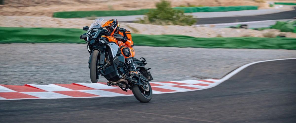 A motorcyclist performing a wheelie on a track, wearing an orange and black racing suit.
