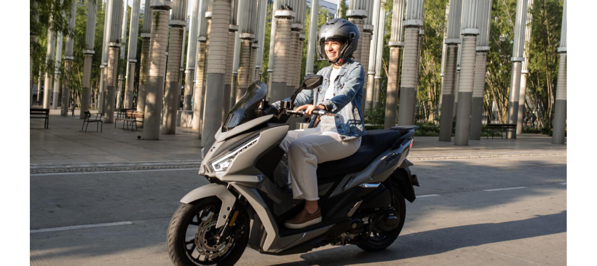 A smiling person wearing a helmet is riding a gray scooter on a city street, surrounded by tall, modern columns.