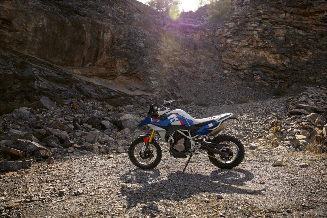 An adventure motorcycle positioned on a rocky terrain with a backdrop of steep cliffs and sunlight filtering through the trees.