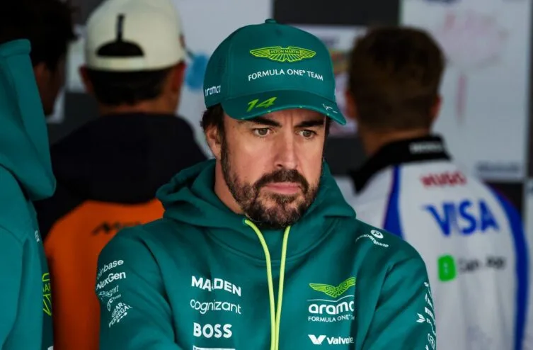 A serious-looking man in a green Aston Martin Formula One hoodie and cap, standing in a paddock environment with blurred figures in the background.