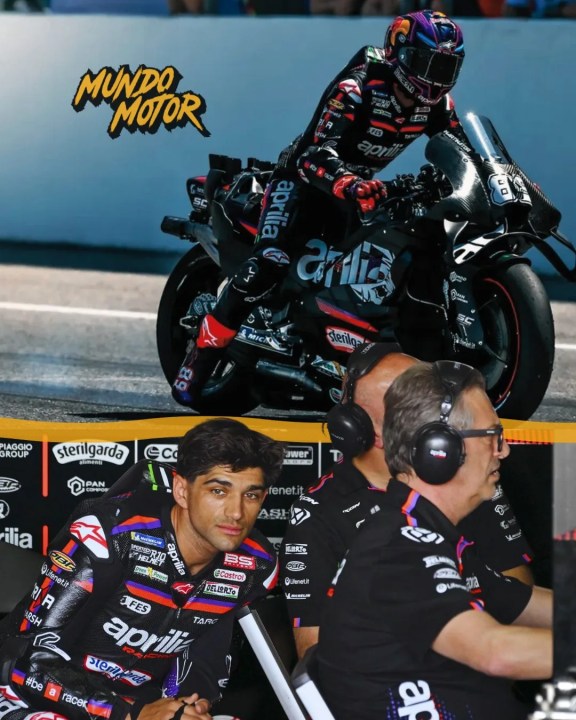 A motorcycle rider in racing gear riding an Aprilia motorcycle on a track, with a pit crew team observing in the background.