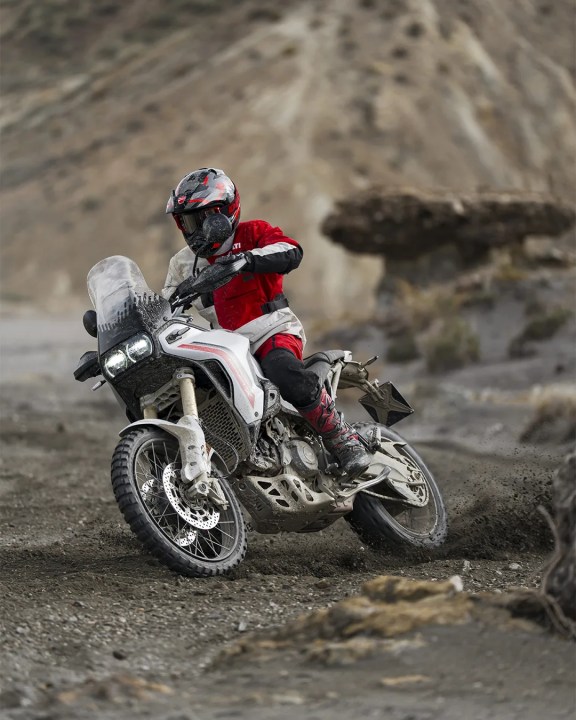 A motorcyclist in red and black gear riding an adventure motorcycle on a rugged dirt path, kicking up dust and gravel.