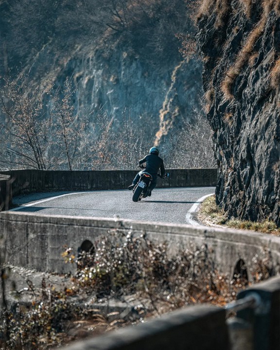 A Ducati Formula 73 (2026) motorcyclist riding along a winding mountain road surrounded by rocky cliffs and sparse trees.
