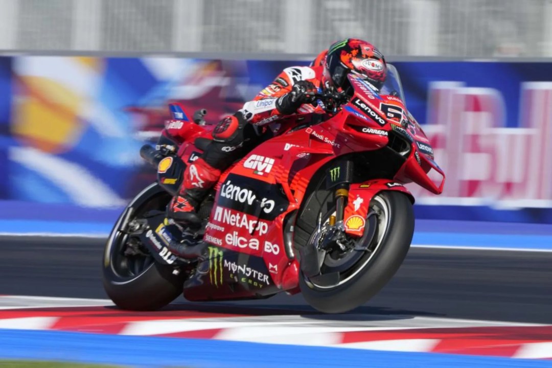 A motorcycle racer in a red racing suit leans into a turn while riding a Ducati motorcycle on a racetrack, with a vibrant background of blue and colorful graphics.