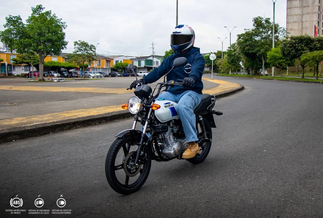 Motociclista con casco y chaqueta oscura, montado en una moto Suzuki, circulando por una calle con edificios y árboles al fondo.