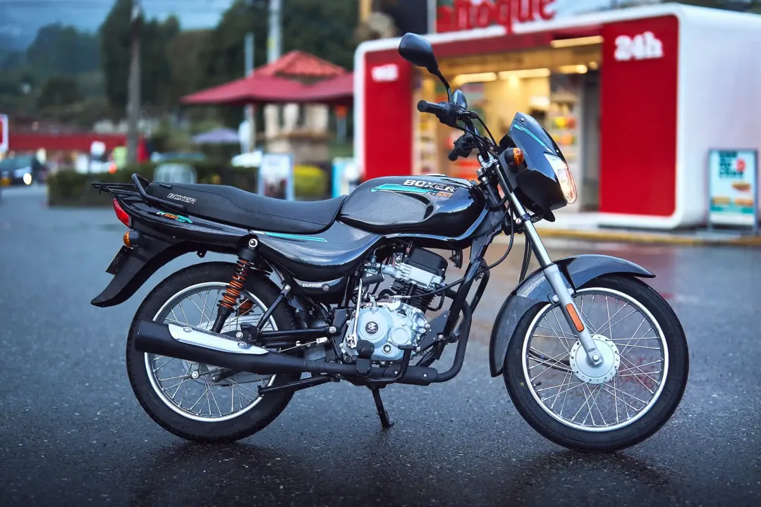 Moto negra modelo Boxer estacionada en una calle con asfalto mojado, fondo difuso de un establecimiento.