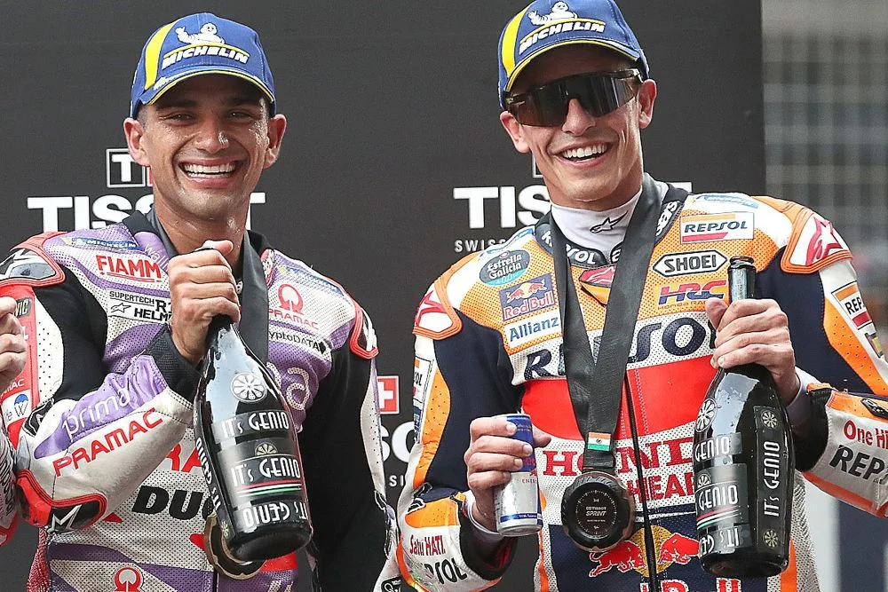 Two motorcycle racers celebrating on the podium, holding trophies and drinks, smiling at the camera.