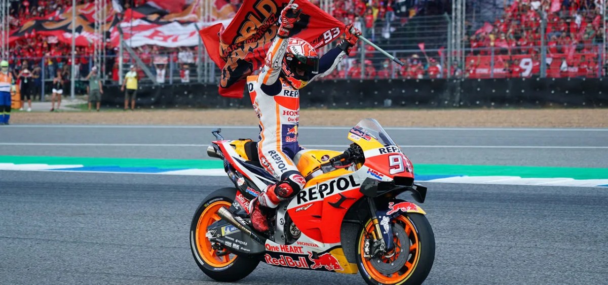 A motorcycle racer celebrating on the track while holding a flag, with a cheering crowd in the background.