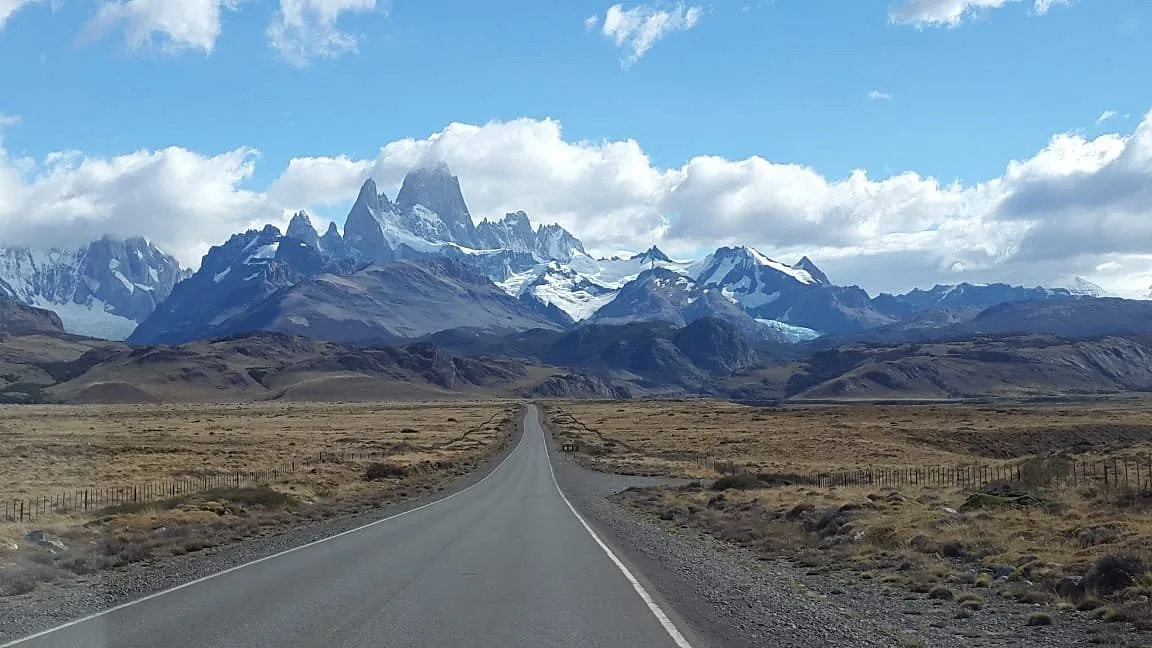Ruta asfaltada rodeada de montañas cubiertas de nieve bajo un cielo azul con nubes.