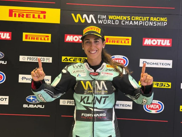 Piloto de motociclismo en su traje de competición, sonriendo y levantando ambos dedos índices en señal de victoria, con un fondo de patrocinadores del campeonato mundial de motociclismo femenino.