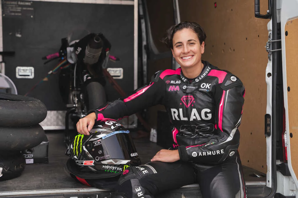 Piloto de motociclismo sonriendo con equipo de carreras negro y rosa, junto a su casco y neumáticos en un remolque.