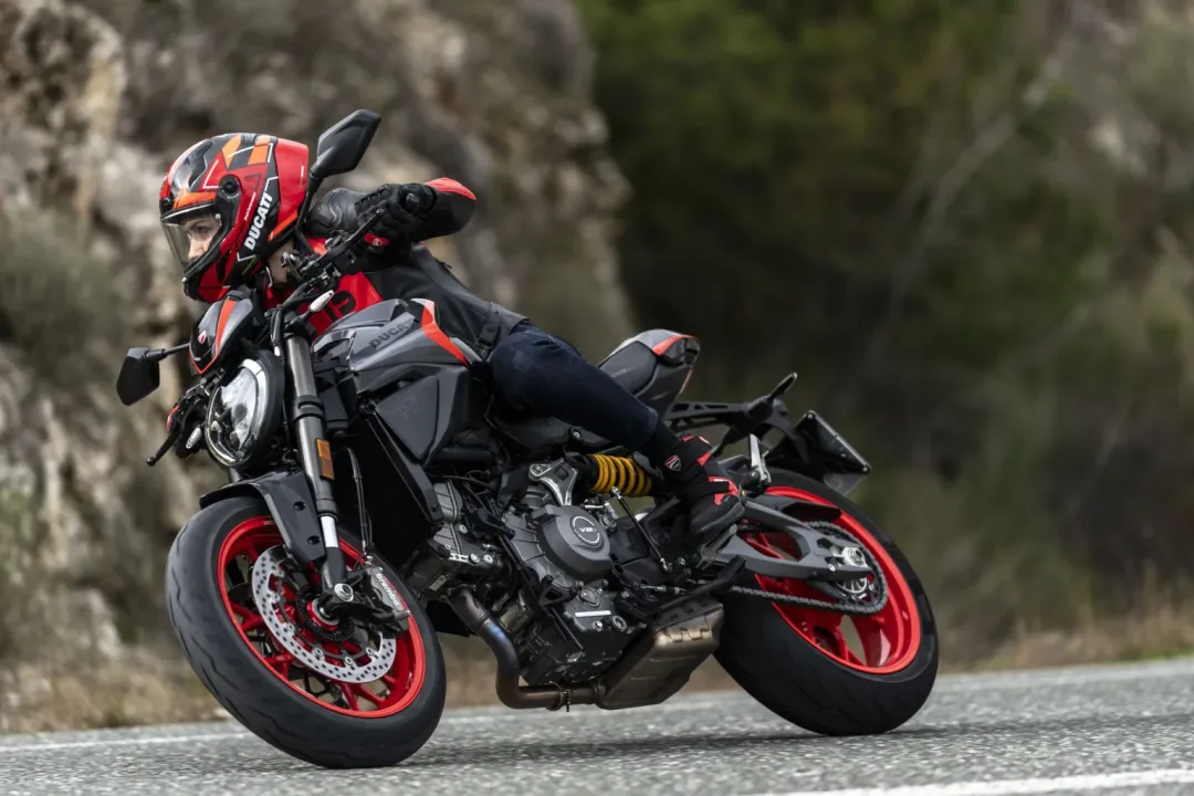 A motorcyclist in riding gear leans into a turn on a Ducati motorcycle, showcasing the bike's sporty design and vibrant red accents against a blurred natural background.