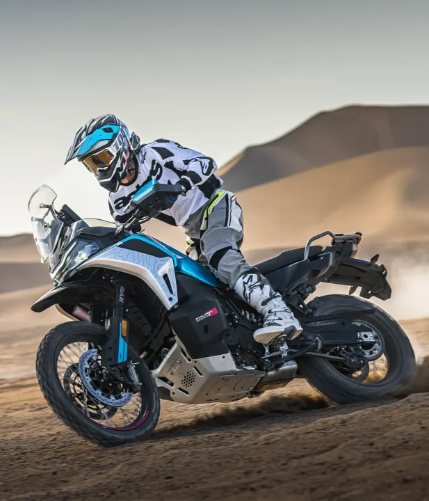 A rider in protective gear skillfully maneuvers a motorcycle on a sandy terrain with desert hills in the background.