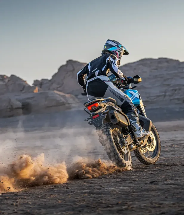 A motorcycle rider kicking up dust while riding on a dirt road in a desert landscape, with rocky formations in the background.