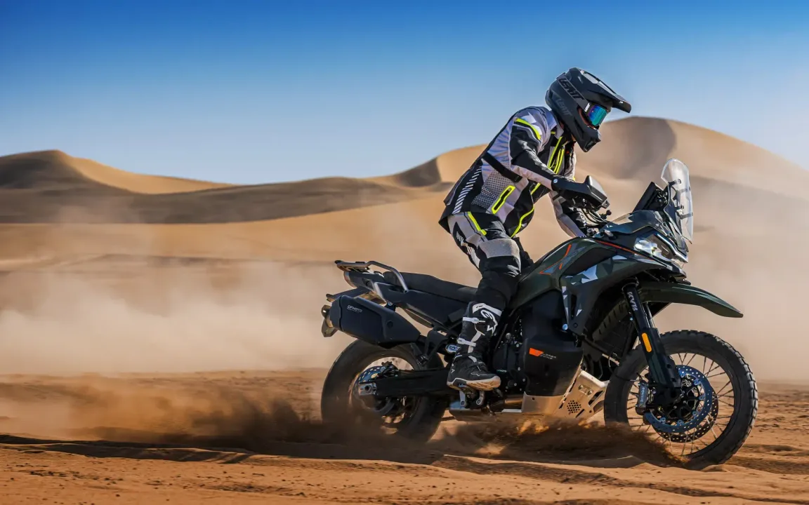 A motorcyclist wearing protective gear rides an off-road motorcycle in a sandy desert landscape, kicking up dust behind them.