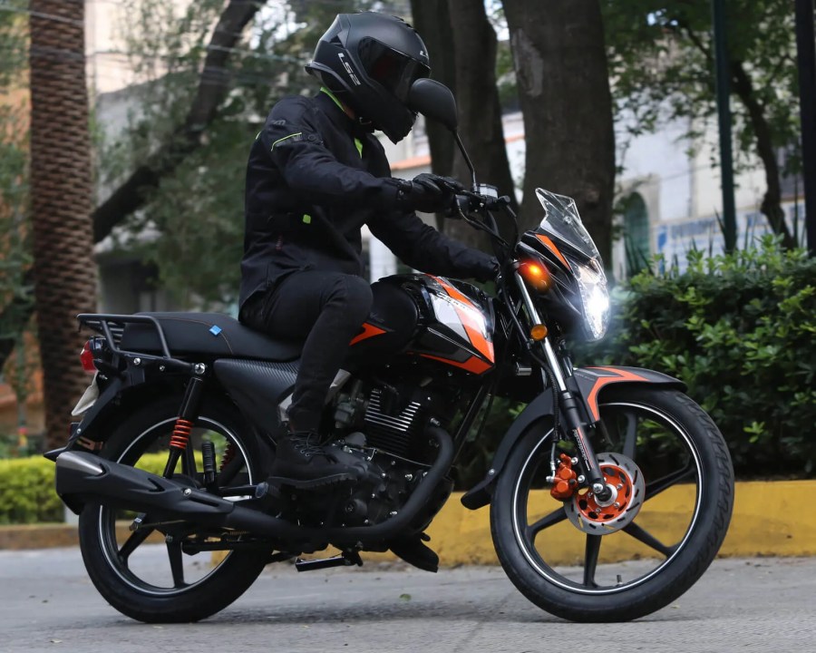 Motociclista con chaqueta negra y casco, montado en una moto de color negro y naranja, circulando por una calle con árboles y arbustos.