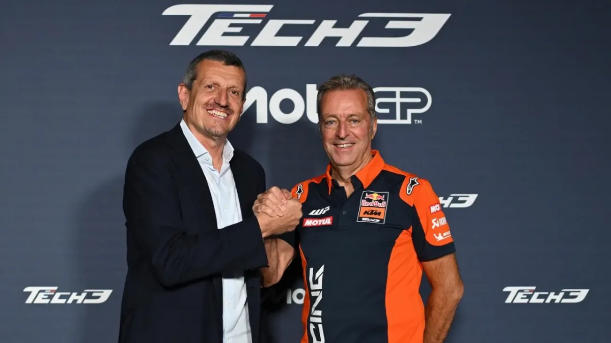 Two men shaking hands in front of a Tech3 logo backdrop, one wearing a black suit and the other in an orange and black racing team uniform.