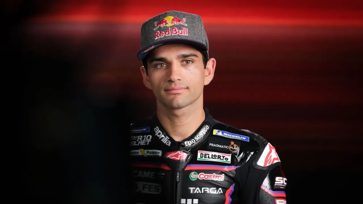 A motorcycle racer wearing a Red Bull cap and a racing suit featuring various sponsor logos, posing against a dramatic red background.