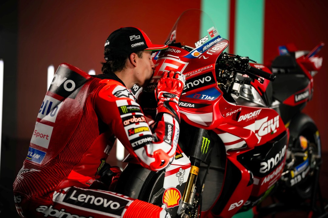 A motorcycle racer in a red and black racing suit affectionately leans in to kiss the front of a Ducati racing motorcycle in a promotional setting.
