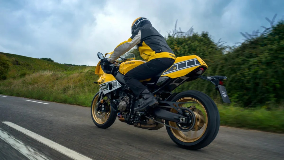 A rider in a yellow and black Yamaha jacket is riding a motorcycle on a rural road, surrounded by greenery and under a cloudy sky.
